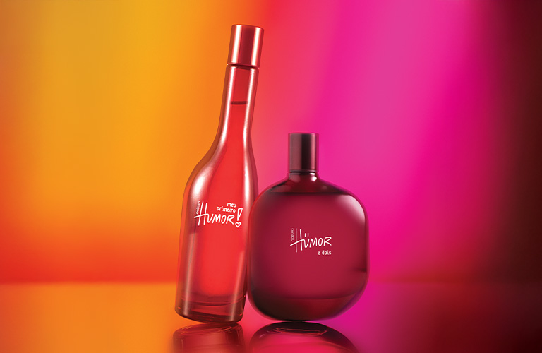 Fotografía de dos frascos de perfume Humor en tonos rojos. El fondo presenta un degradado vibrante en colores naranja, rosa y amarillo.